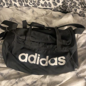 Svart träningsväska från Adidas - Snygg svart träningsväska från Adidas med stort vitt logotryck på sidan. Väskan har både handtag och justerbar axelrem, samt dragkedja upptill. Perfekt storlek för gymmet eller sportaktiviteter. Tillverkad i slitstark polyester.