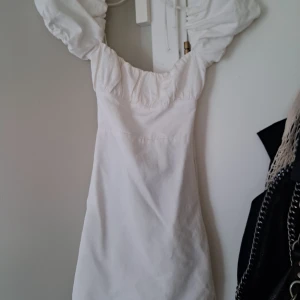 Vit kort klänning med puffärmar från Zara - Supersöt vit kort klänning från Zara med puffiga kortärmar och öppen rygg med snörning. Klänningen har en figurnära passform och är gjord i mjukt bomullstyg. Perfekt för sommarens alla tillfällen när du vill känna dig extra fin.