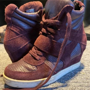 Vinröda wedge sneakers med mocka - Säljer ett par vinröda sneakers med inbyggd kilklack och snygga detaljer i mocka och perforerat syntetmaterial. Skorna har snörning och kardborreband för extra cool look. Perfekt för dig som vill ha lite höjd men ändå känna dig bekväm.