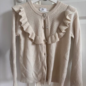 Beige kofta med volang från Noa Noa - Supersöt beige kofta från Noa Noa i stickad kvalitet med pärlknappar och volangdetaljer längs axlar och framstycke. Framsidan har ett snyggt strukturerat mönster och koftan är långärmad med rund hals. 