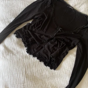 Svart croppad blus med spets Gina Tricot - Supersöt svart croppad blus från Gina Tricot i storlek S. Toppen har långa ärmar, spetsdetaljer längs halsringningen och framtill, liten rosett och volangkant nedtill. Perfekt för dig som gillar romantiska och trendiga vibes.  