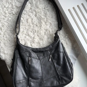 Svart axelväska  - Väska från Verona, har fickor både fram och baksida. Den har två större fack och i varje fack finns de en mindre ficka på insidan av väskan. Justerbar axelband.