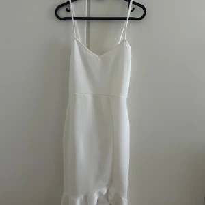 Vit omlottklänning med volang - Säljer en stilren vit kortklänning med smala axelband och v-ringad hals. Klänningen har en omlott-design och en snygg volangdetalj längst ner. Perfekt för sommarens alla tillfällen, enkel att matcha med accessoarer.