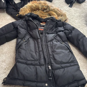 Svart Parajumpers dunjacka med päls - Säljer en svart Parajumpers Anchorage dunjacka med fluffig päls på huvan och snygga detaljer som patch på ärmen. Jackan har långa ärmar, dragkedja och stora fickor framtill. Perfekt för kalla dagar och riktigt stilren look.