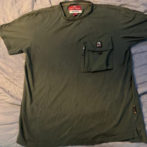 Grön t-shirt Parajumpers S med ficka - Snygg grön t-shirt från Parajumpers i storlek S. T-shirten har en stor bröstficka med lock, dragkedja och coolt PJS-märke. Materialet är mjukt och skönt fleece, perfekt för chill dagar. Passformen är normal och ärmarna är korta.