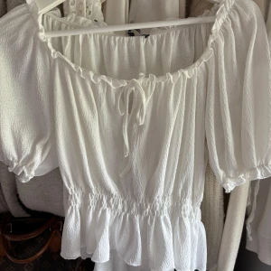 Vit blus med volang från Gina Tricot - Supersöt vit blus från Gina Tricot med puffiga korta ärmar, volangkant nedtill och smock i midjan. Den är helt ny och oanvänd