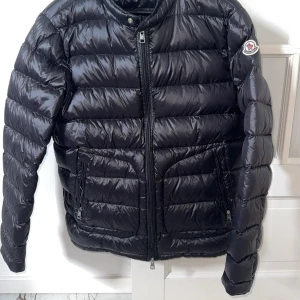 Svart Moncler pufferjacka herr - Svart pufferjacka från Moncler med glansig finish och klassisk logga på ärmen. Jackan har två sidofickor med dragkedja, ståkrage och quiltad design. Perfekt för kalla dagar och har snygga detaljer som dubbla dragkedjor framtill. Storlek M/ liten L