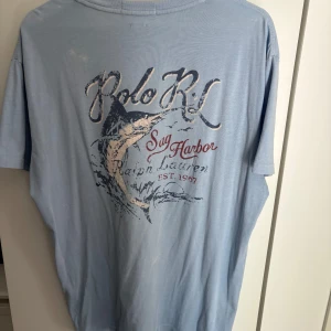 Ljusblå Polo Ralph Lauren t-shirt - Snygg ljusblå t-shirt från Polo Ralph Lauren med tryck av en svärdfisk och texten 'Sag Harbor' på bröstet. Klassisk rund hals och korta ärmar. Materialet är mjuk bomull, perfekt för chill dagar. Trycket har en cool vintage-look med inslag av marinblått och rött.