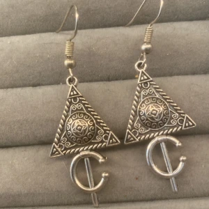 Silvriga triangelformade örhängen - Unika örhängen i silverfärgad metall med triangelform och detaljerade mönster. Framsidan har ett cirkulärt ornament med snirkliga linjer och en stjärnliknande design i mitten. Nedanför hänger en rundad symbol som ger en cool, bohemisk vibe.