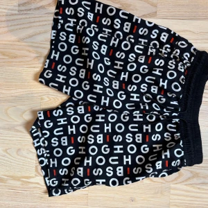 Svarta shorts från Hugo Boss XS - Snygga svarta shorts från Hugo Boss i storlek XS med elastisk midja. Shortsen har ett coolt grafiskt mönster med vita och röda bokstäver över hela tyget. Materialet är mjukt och lätt, perfekt för varma dagar. Passformen är loose och de har fickor på sidorna.