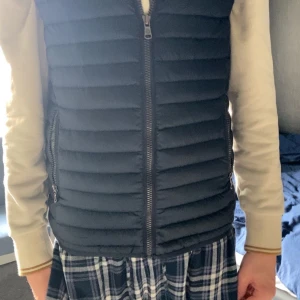 Svart dunväst med dragkedja - Säljer en svart dunväst med horisontella quiltade paneler och två sidofickor med dragkedja. Västen har en rak passform och hel dragkedja framtill. Perfekt att slänga över en hoodie eller tröja när det är kyligt ute.