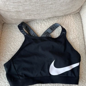 Svart Nike sporttopp med logoband - Sportig svart topp från Nike med breda, korsade axelband där det står 'NIKE' i vitt. Stort vitt swoosh-tryck framtill och tillverkad i Dri-FIT polyester som andas. Snygg croppad passform och perfekt för träning eller gymmet.