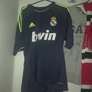 Real Madrid Ronaldo 7 marinblå matchtröja - Säljer en officiell Real Madrid matchtröja från Adidas med Ronaldo 7 på ryggen. Marinblå med neongula detaljer och klubbmärke, LFP-patch på ärmen och jubileumsmärke. Tillverkad i lätt och ventilerande polyester med ClimaCool-teknologi. Perfekt för dig som älskar fotboll och Cristiano Ronaldo.