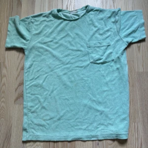 Mintgrön frotté t-shirt med ficka - Fräsch mintgrön t-shirt i mjuk frotté med rund hals och en praktisk bröstficka. Modellen är rak och har korta ärmar, perfekt för chill dagar. Materialet är skönt och ger en avslappnad vibe. Enkelt och stilrent plagg som funkar till jeans eller shorts.