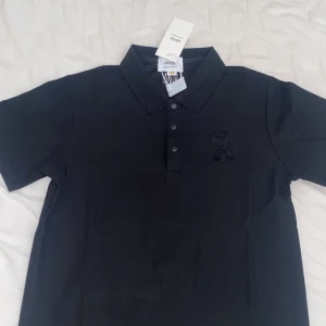 Svart polo t-shirt från AMI - Svart polo t-shirt från AMI med klassisk krage och tre knappar framtill. På bröstet finns ett snyggt broderat hjärta med bokstaven A. Materialet är mjuk bomull och passformen är slim. AMI-logga broderad i nacken för extra stilpoäng.