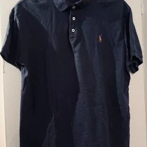 Mörkblå polo t-shirt från Ralph Lauren - Snygg mörkblå polo t-shirt från Ralph Lauren med klassisk krage och tre vita knappar framtill. Ikoniska broderade loggan på bröstet. Perfekt för dig som gillar stilrena och tidlösa plagg. Skön bomullskvalitet och korta ärmar.
