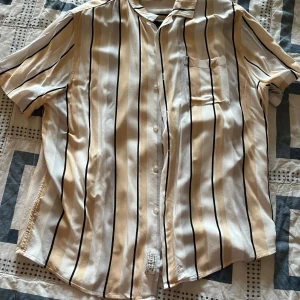 Randig kortärmad skjorta från Pier one - Snygg kortärmad skjorta från pier one med vertikala svarta och beige ränder på vit botten. Skjortan har klassisk krage, bröstficka och knappar framtill. Perfekt för chill dagar och sommarhäng.