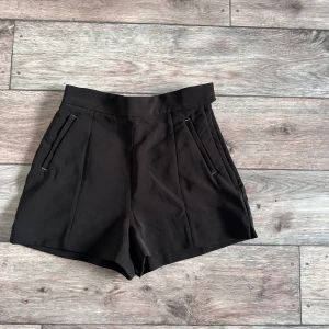 Svarta högmidjade shorts från H&M - Stilrena svarta shorts från H&M med medelhög midja och raka ben. De har två fickor fram och dekorativa fickor bak. Snygg, enkel design som passar perfekt till en trendig look. Gjorda i ett mjukt och lätt material som känns bekvämt hela dagen.