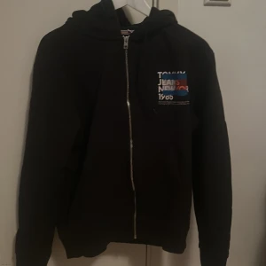 Svart hoodie Tommy Jeans med tryck - Svart hoodie från Tommy Jeans med stort tryck i vitt, blått och rött på ryggen och mindre tryck på bröstet. Tröjan har huva, dragkedja och långa ärmar. Perfekt för dig som gillar streetwear och vill ha en snygg och bekväm look.