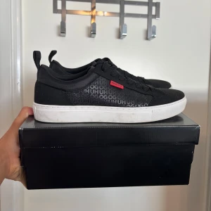 Svarta skor från Hugo Boss - Stilrena svarta skor från Hugo Boss med vit sula och diskret logomönster på sidan. Skorna har en  röd Hugo-detalj på sidan. Perfekta för en clean och modern look.