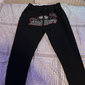 Svarta mjukisbyxor med tryck från trapstars unika plagg  - Svarta mjukisbyxor med coolt tryck framtill där det står 'Shooters' och har en pantergrafik. Byxorna har resår och snörning i midjan för skön passform. Perfekta för chill eller träning.