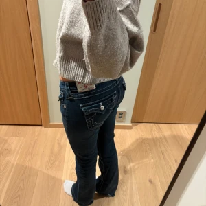 True Religion mörkblå jeans bootcut - Snygga mörkblå jeans från True Religion med bootcut och kontrastsömmar. Jeansen har låg midja och ”krystal” knappar. Säljes billigt pga slitage längst ner på byxorna.