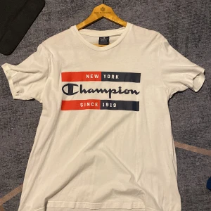 Vit Champion t-shirt med tryck - Snygg vit t-shirt från Champion med stort tryck på bröstet i rött, blått och vitt. Klassisk rund hals och korta ärmar. Perfekt för dig som gillar streetwear och vill ha en enkel men stilren look. Tillverkad i mjuk bomull för skön känsla hela dagen.