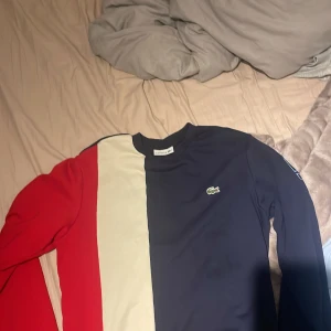 Flerfärgad långärmad tröja Lacoste - Säljer en snygg långärmad tröja från Lacoste med breda block i rött, vitt och mörkblått. Tröjan har klassisk rund hals och den ikoniska krokodilloggan på bröstet. Perfekt för dig som gillar sportig och stilren design.
