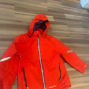 Röd Everest vindjacka med huva - Riktigt snygg röd vindjacka från Everest med huva och dragkedja framtill. Jackan har svarta detaljer, reflex på ärmen och två dragkedjeförsedda fickor. Perfekt för blåsiga dagar och outdoor-äventyr. Sportig look och skön passform.