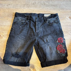 Svarta jeansshorts med rosbrodyr - Svarta jeansshorts från Denim Co. i slim fit med snygga slitningar och uppvikta benslut. På ena benet finns en broderad detalj med röda rosor och gröna blad som ger en cool touch. Klassisk femficksmodell och knappgylf. Perfekta för dig som gillar streetstyle.