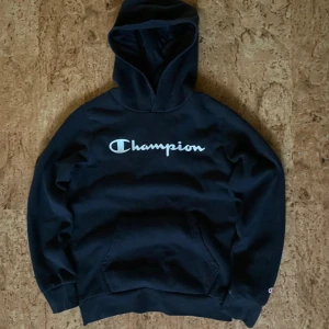 Svart Champion hoodie med logga - Svart hoodie från Champion med stor vit logga över bröstet. Klassisk känguruficka framtill och huva. Mjuk insida och ribbade muddar vid ärmslut och nederkant. Perfekt för en chill och sportig stil.