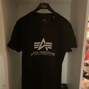 Svart Alpha Industries t-shirt - Svart t-shirt från Alpha Industries med stor vit logga och text på bröstet. Klassisk rund hals och korta ärmar. Perfekt för dig som gillar streetwear och vill ha en clean look med tydlig branding.