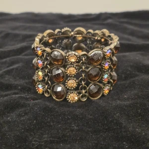 Brett armband med bruna stenar - Unikt brett armband med stora bruna och guldiga stenar i flera rader. Armbandet har antikguld-färgad metall och gnistrande detaljer som ger en cool vintagekänsla. Perfekt för dig som vill sticka ut med accessoarer som verkligen syns.