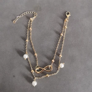 Vintage stil fotlänk  - Detta är ett glittrande fotlänk med 2 st kedjor som sitter ihop. Den ena kedjan är dekorerad med en infinity symbol och blanka guld kulor. Den andra kedjan är dekorerad med vita pärlor och blanka guld kulor. Materialet är copper alloy + simulerad pärlor. Ankellängd är ca 20cm + 5cm. (264)