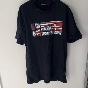 Svart t-shirt från Napapijri - Svart t-shirt från Napapijri med tryck av flera flaggor och logga på bröstet. Klassisk rund hals och korta ärmar. Skön passform och tillverkad i mjuk bomull, perfekt till jeans eller shorts.