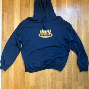 Blå hoodie från Sweet Skateboards - Mörkblå hoodie från Sweet Skateboards med fet logga och flames på bröstet. Klassisk känguruficka, huva med snörning och skön passform. Tillverkad i 100% bomull, perfekt för chill eller skate.