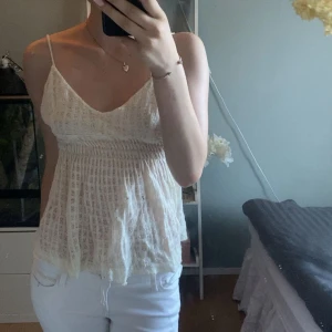 Vit linnetopp från Camilla Norrback - Supersöt vit linnetopp med smala axelband och v-ringning från Camilla Norrback. Toppen är stickad i ett luftigt mönster, har smockad midja och flowig passform. Perfekt för varma dagar och ger en romantisk vibe.