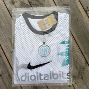 Inter Milan vit bortatröja Nike - Inter Milan bortatröja från Nike i vitt med svarta och turkosa detaljer. Tröjan har korta ärmar, svart krage och coolt grafiskt mönster med ormar och växter. Digitalbits sponsortryck på bröstet och klubbmärke i turkos. Perfekt för dig som vill sticka ut på planen. Pris kan diskuteras vid snabb affär 