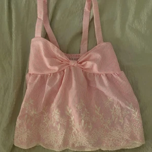 Gullig topp - Supersöt rosa och vit randig linnetopp med breda axelband och stor rosett framtill. Toppen har broderade blommor längs nederkanten och smockad rygg för extra komfort. Perfekt för varma dagar och ger en gullig, somrig vibe.