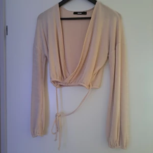 Beige omlottopp från Bik Bok - Säljer en beige omlottopp från Bik Bok med långa ärmar och knytband i midjan. Toppen är croppad och har en djup v-ringning som ger en snygg och trendig look. Perfekt att styla med höga jeans eller kjol.