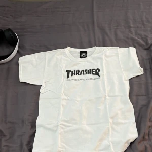 Vit Thrasher t-shirt med tryck - Vit t-shirt från Thrasher med klassiskt svart logotryck på bröstet. Skön passform och tillverkad i mjuk bomull. Perfekt för dig som vill ha en clean look med streetkänsla.