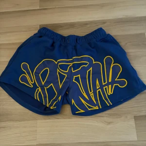 Blå shorts med gul graffiti-tryck - Snygga blå shorts med elastisk midja och stort gult graffiti-inspirerat tryck framtill. Modellen är loose fit och har en bakficka med dragkedja. Perfekta för dig som gillar streetstyle och vill sticka ut med färg och mönster.