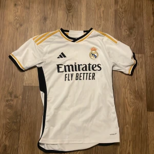 Real Madrid Adidas vit matchtröja - Snygg Real Madrid fotbollströja i vitt med svarta och gula detaljer. Adidas-logga på bröstet, klubbmärke och 'Emirates Fly Better' tryckt framtill. Klassisk passform med korta ärmar och svarta paneler på sidorna. Tillverkad i lätt och ventilerande material. Jag köpte den på unisports.store för 1600