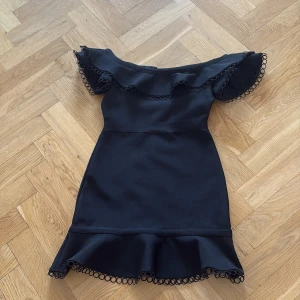 Svart offshoulder klänning Lipsy - Supersnygg svart kortklänning från Lipsy med offshoulder-modell och volangdetaljer vid axlar och fåll. Klänningen har dekorativa cirkelkanter och figurnära passform. Perfekt för dig som vill sticka ut med en elegant och trendig look.