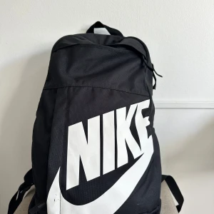 Svart ryggsäck från Nike - Svart ryggsäck från Nike med stor vit logga framtill. Väskan har flera fack med dragkedja, justerbara och vadderade axelremmar samt ett praktiskt bärhandtag upptill. Tillverkad i slitstark polyester, perfekt för skola eller träning.