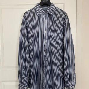 Randig skjorta från Polo Ralph Lauren - Snygg skjorta från Polo Ralph Lauren i blått och vitt med klassiskt randigt mönster. Skjortan är tillverkad i 100% bomull, har lång ärm, bröstficka och knäppning framtill. Perfekt för dig som gillar stilrena och tidlösa plagg.