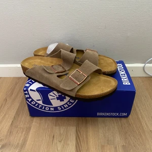 Birkenstock Arizona - Snygga sandaler från Birkenstock i beige mocka med två breda remmar och justerbara spännen i kopparfärg. Fotbädden är mjuk och anatomiskt utformad för extra komfort. Perfekta för varma dagar och chill vibes.