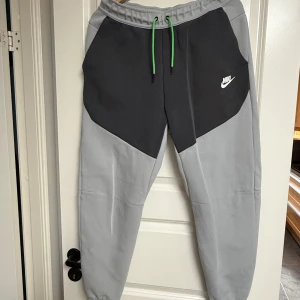 Grå Nike Tech Byxor - Snygga Nike Tech mjukisbyxor från Nike i två nyanser av grått med coola gröna snören i midjan. Byxorna har elastisk midja, sidofickor och en vit Nike-logga på låret. Perfekta för chill eller träning, tillverkade i mjukt bomullsmaterial.