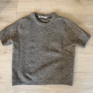 Grå stickad tröja från Zara - Mysig grå stickad tröja från Zara med korta ärmar och rund hals. Tröjan har ribbade muddar vid ärmslut och nederkant, perfekt för lager-på-lager och chill vibes. Enkel och stilren design som funkar till det mesta.