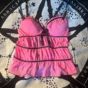 Rosa korsettopp med volanger och rosett - Supersöt rosa korsettopp med svarta detaljer och volanger. Toppen har vadderade kupor, justerbara axelband och en svart rosett framtill. Baksidan har hysk- och hakknäppning. Perfekt för dig som vill sticka ut med en feminin och lekfull stil.